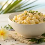 A large white ceramic bowl filled with creamy Hawaiian macaroni salad, set on a light wooden table with tropical leaves in the background.